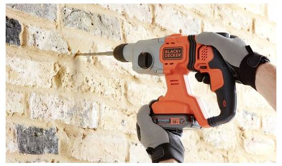 Eine Person, die Handschuhe trägt, benutzt einen orange-schwarzen Elektroschrauber, um ein Loch in eine Backsteinmauer zu bohren.