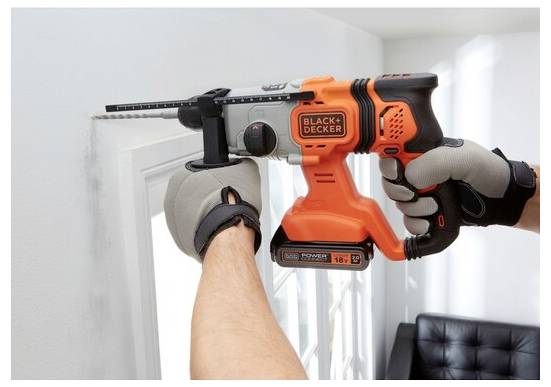 Eine Person benutzt einen orange-schwarzen kabellosen Elektroschrauber, um ein Loch in eine Wand neben einem Fenster zu bohren. Die Hände tragen Schutzhandschuhe.