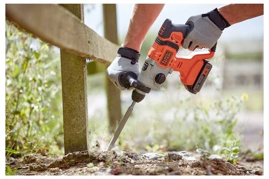 Eine Person in Handschuhen verwendet einen orange-schwarzen Elektroschrauber, um einen Holzpfosten im Boden zu befestigen, umgeben von Gras und Sonnenlicht.