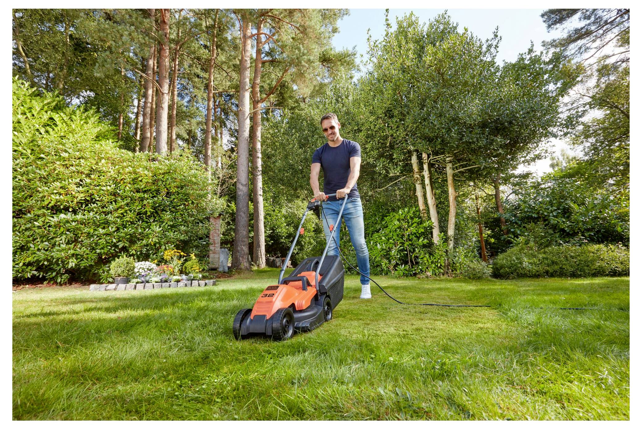 Eine Person mäht einen üppig grünen Rasen mit einem elektrischen Rasenmäher in einem Garten, der von hohen Bäumen und Sträuchern umgeben ist.