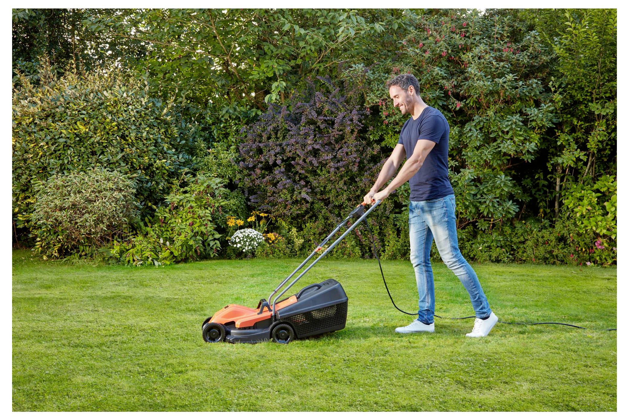 Eine Person mäht einen grünen Rasen mit einem elektrischen Rasenmäher in einem Garten, umgeben von Büschen und Bäumen unter einem klaren Himmel.