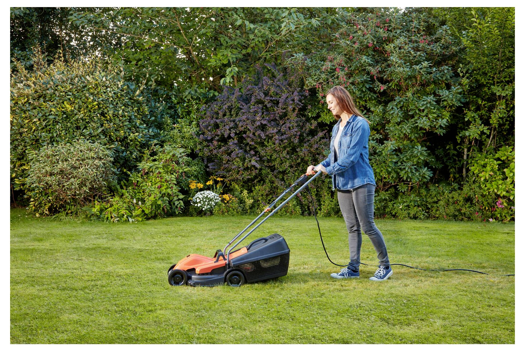 Eine Person in legerer Kleidung benutzt einen orangefarbenen elektrischen Rasenmäher, um Gras in einem Garten zu schneiden, der von grünen Sträuchern und Bäumen umgeben ist.