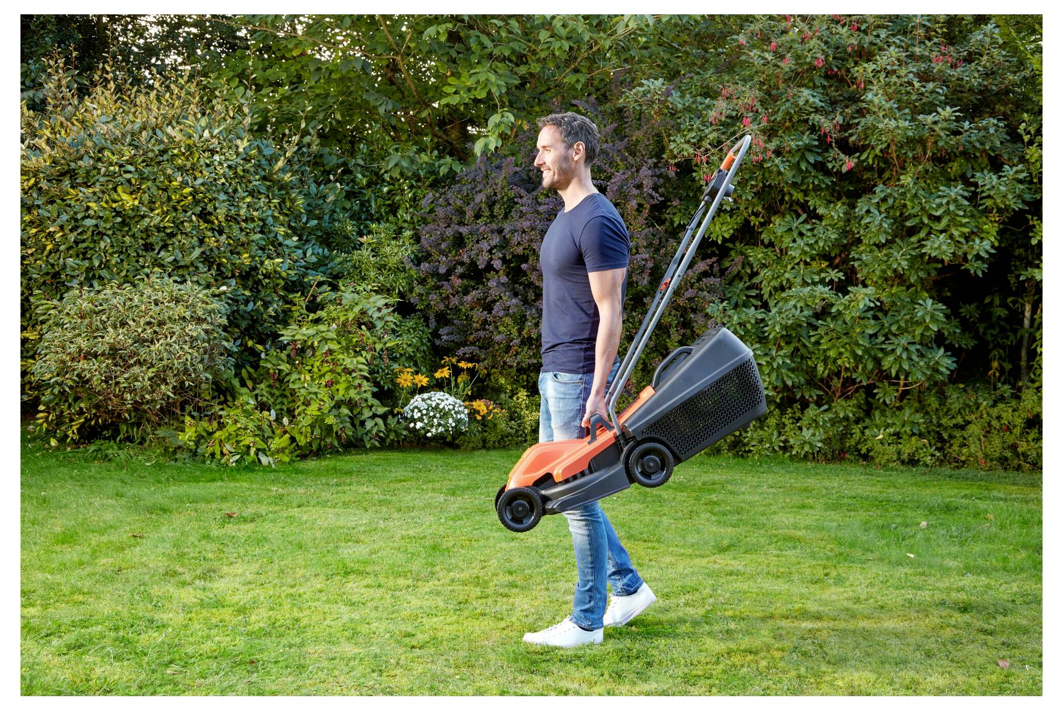 Eine Person in einem Garten trägt einen kleinen Rasenmäher. Das Gras ist ordentlich geschnitten, und grüne Büsche mit Blumen sind im Hintergrund.