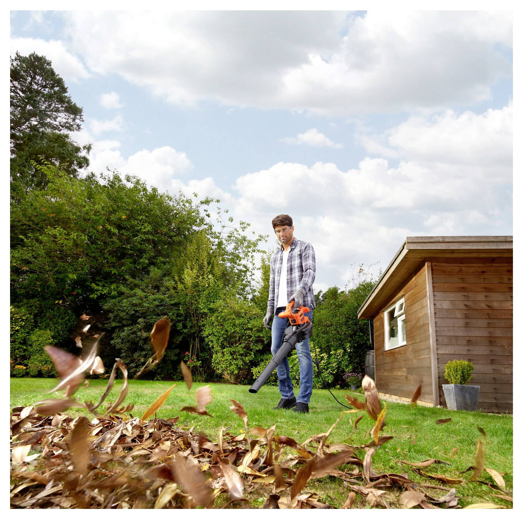 Eine Person benutzt einen Laubblasgerät, um gefallenes Laub in einem Garten zu entfernen, mit einem hölzernen Schuppen und Bäumen im Hintergrund an einem sonnigen Tag.