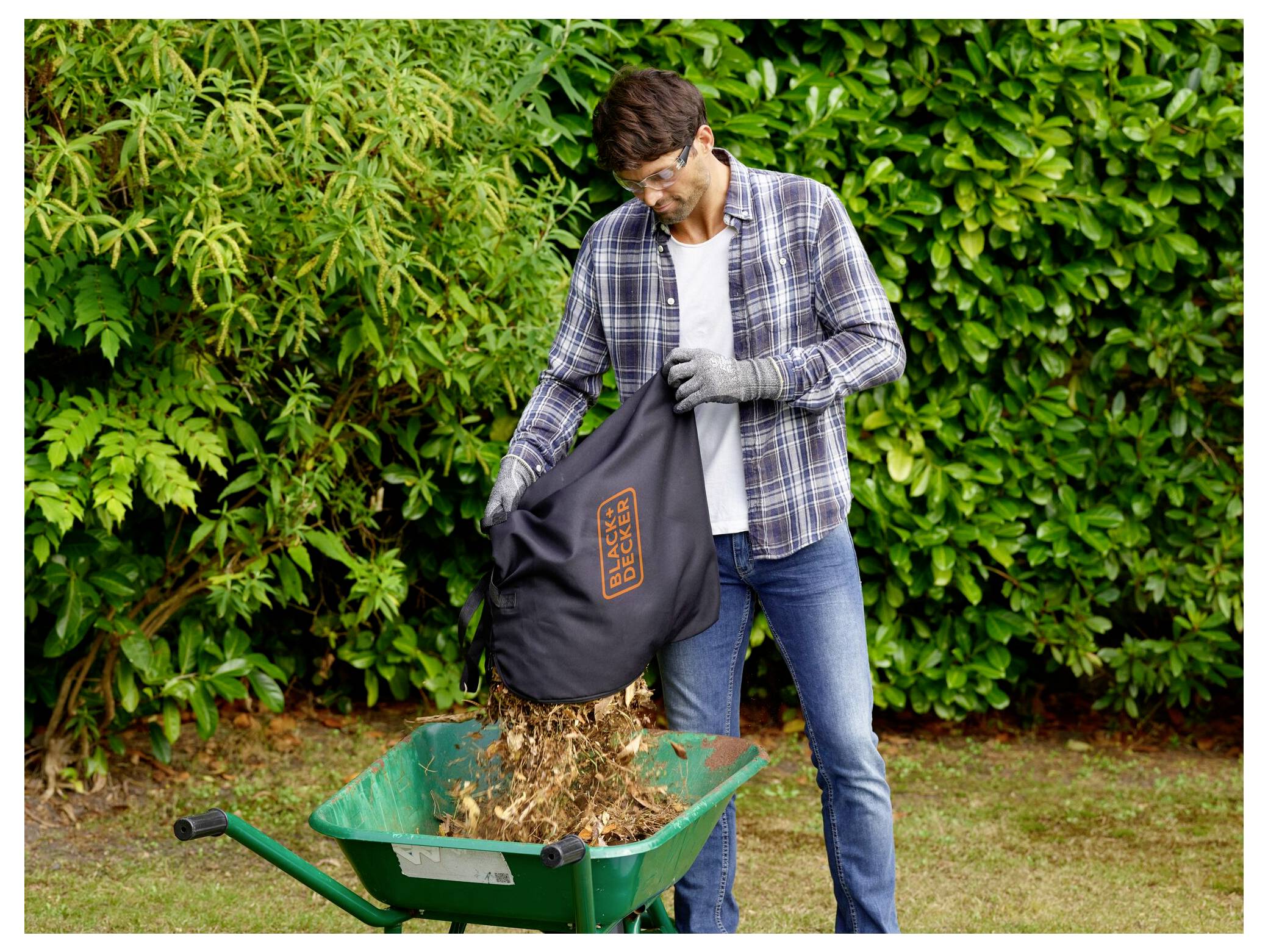 Eine Person mit Handschuhen gießt Gartenabfälle aus einem Sack in eine grüne Schubkarre und steht dabei vor üppig grünen Büschen.