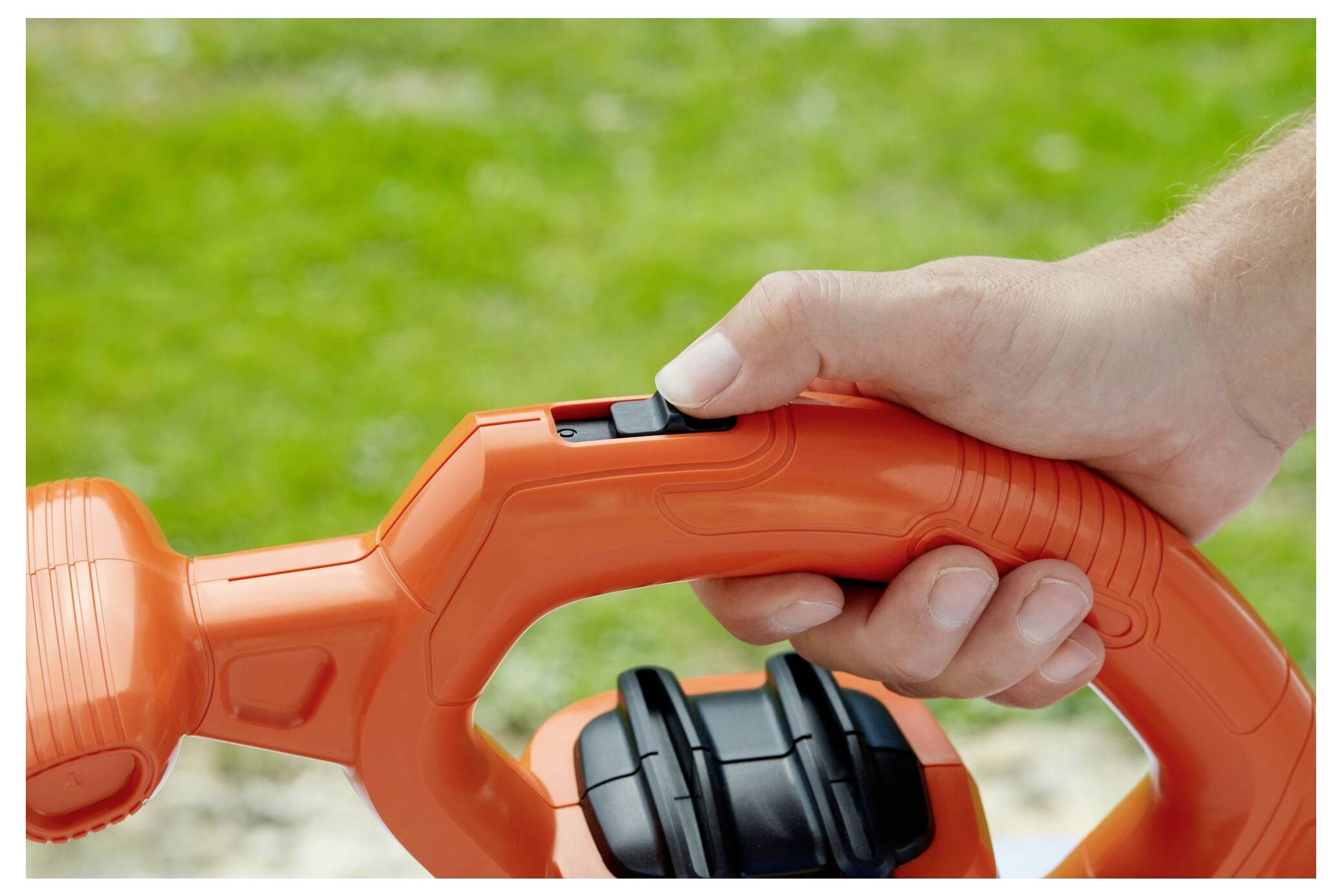 Eine Person hält den Griff einer orangefarbenen Gartenschere in der Hand, drückt einen schwarzen Knopf, vor einem grünen Grashintergrund.