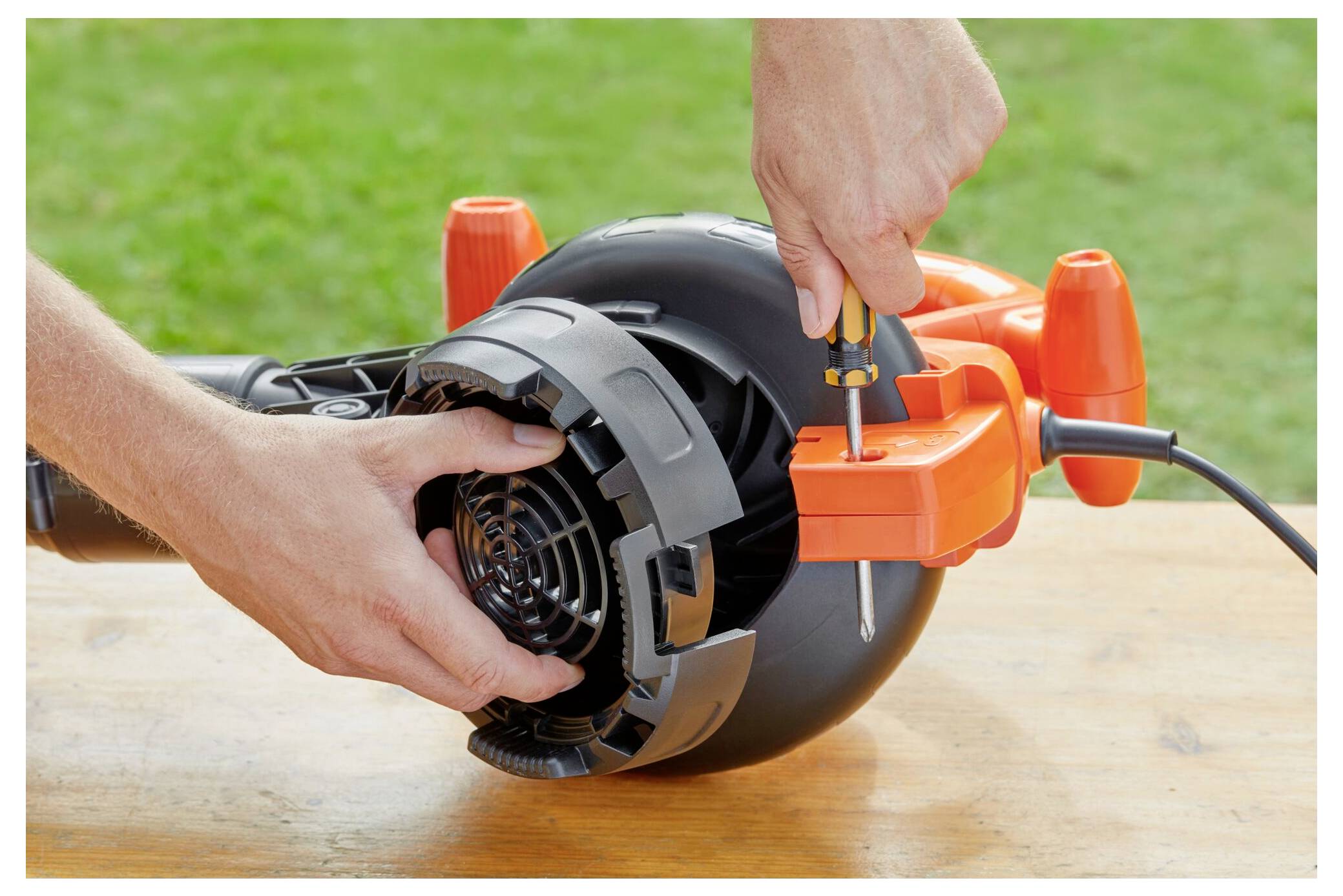 Eine Person verwendet einen Schraubendreher, um ein orange-schwarzes Elektrowerkzeug auf einer Holzoberfläche zusammenzubauen oder zu reparieren, wobei Gras im Hintergrund sichtbar ist.