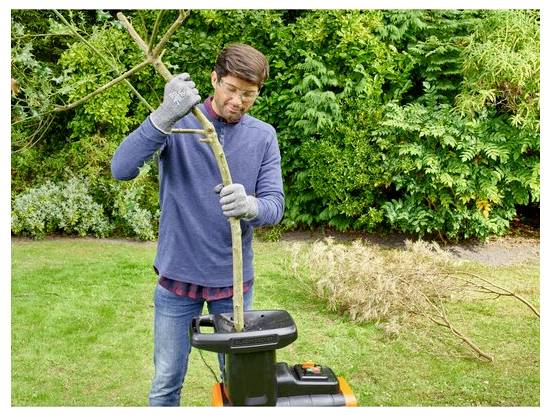 Eine Person bedient einen Gartenhäcksler und führt einen Baumast in die Maschine ein, mit dichtem Grün im Hintergrund.