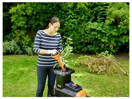 Eine Frau in einem gestreiften Hemd benutzt einen elektrischen Gartenhäcksler für Äste in einem Garten, umgeben von üppigen grünen Bäumen und Gras.