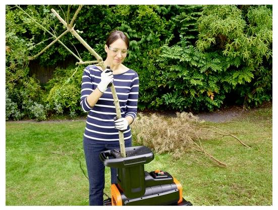 Eine Person in einer Schutzbrille und Handschuhen führt Äste in einen Gartenhäcksler ein, mit Grünflächenresten und getrockneten Ästen im Hintergrund.