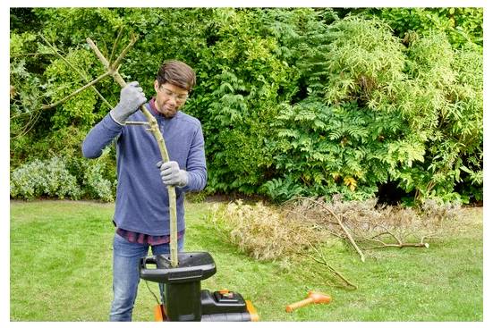 Ein Mann mit Handschuhen führt Äste in einen Gartenhäcksler ein, umgeben von Grünflächen und geschnittenen Ästen in einer Hinterhof-Umgebung.