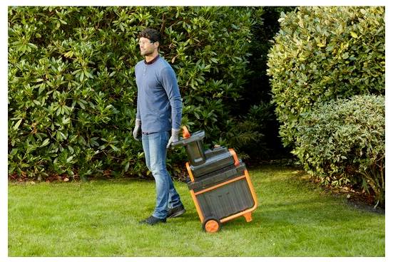 Eine Person in Freizeitkleidung und Handschuhen zieht einen Laubsauger über eine mit dichtem Buschwerk umgebene Grünfläche.