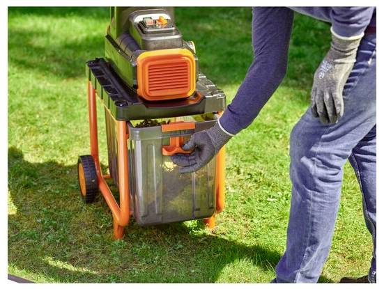 Eine Person trägt Handschuhe und bedient einen orange-schwarzen Laubzerkleinerer auf Gras, was auf die Verwendung für Gartenarbeit und Abfallwirtschaft hindeutet.