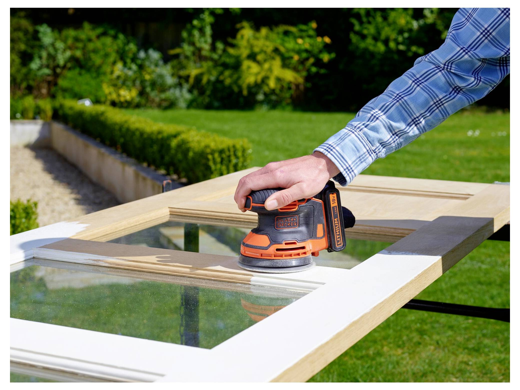 Eine Person schleidet eine Holztür mit einem Elektroschleiffer in einer Gartenumgebung, umgeben von Grünflächern.