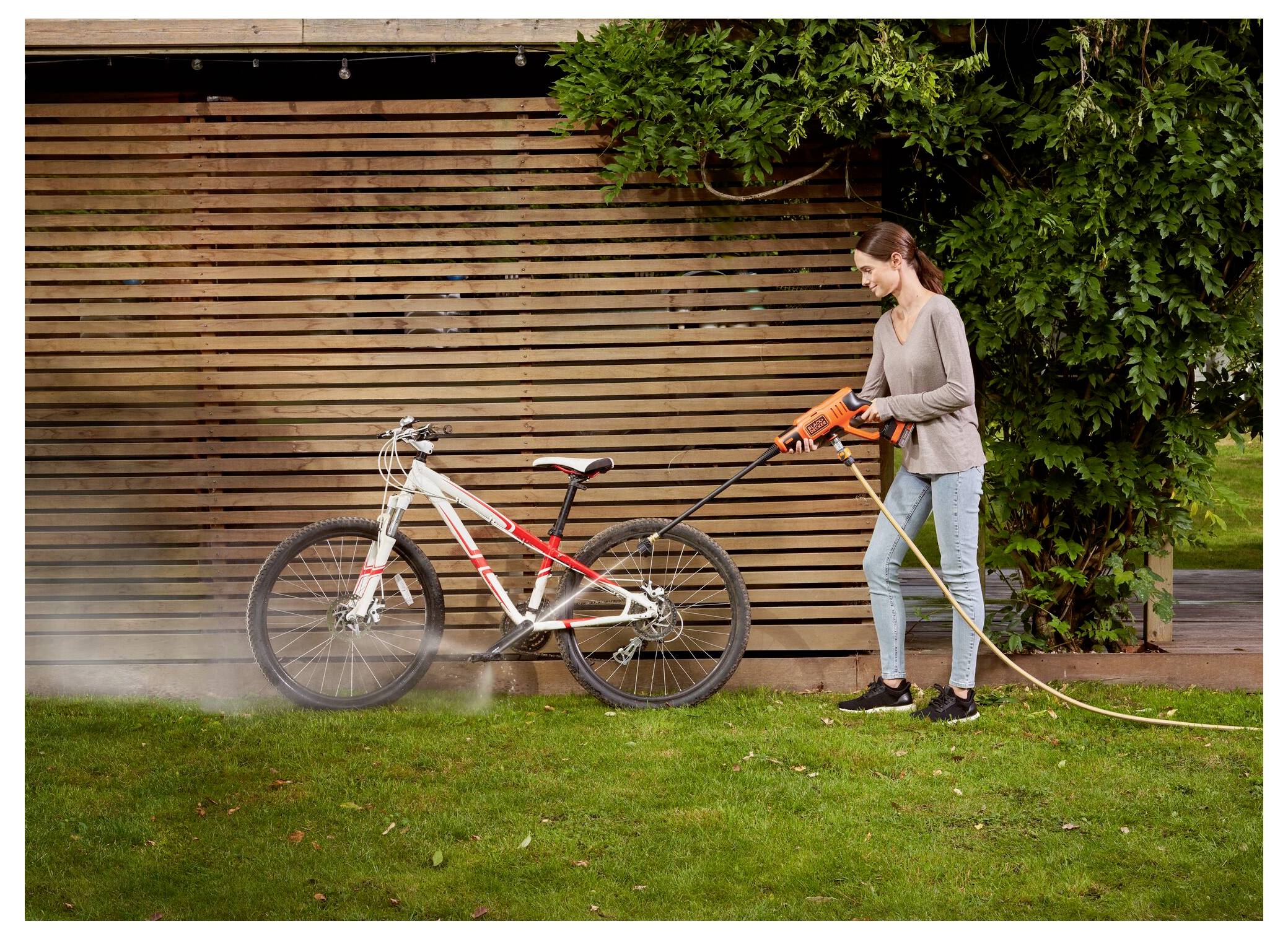 Eine Person wäscht mit einem Schlauch ein Fahrrad auf einem Rasen, mit einem Holzzaun und Grünflächen im Hintergrund.