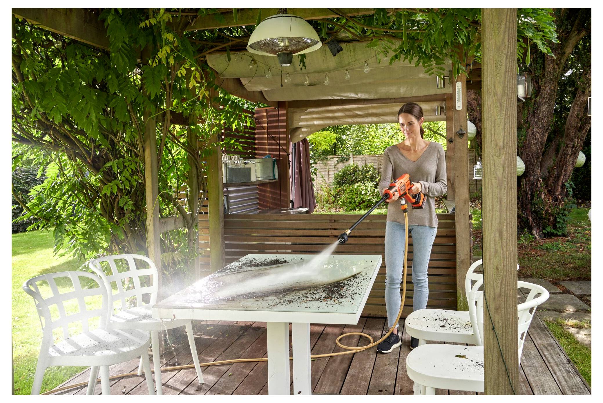 Eine Person benutzt einen Hochdruckreiniger, um einen Außenholztisch auf einer Terrasse zu reinigen, die von Grünflächen umgeben ist, mit Stühlen und einer Pergola in der Nähe.