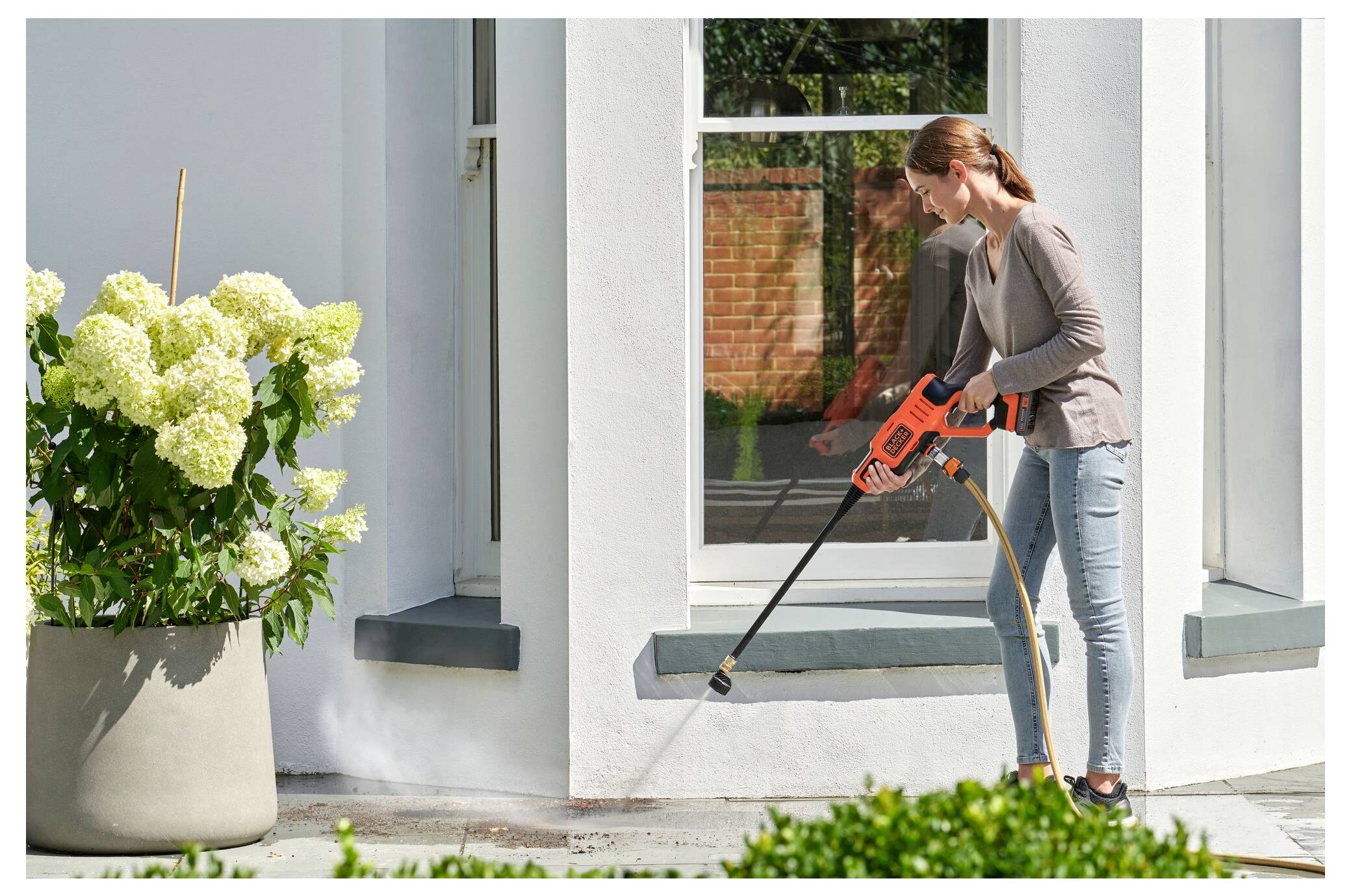 Frau reinigt Terrasse mit Hochdruckreiniger in der Nähe von Blumentöpfen, trägt Freizeitkleidung; reflektierte Glastür im Hintergrund.