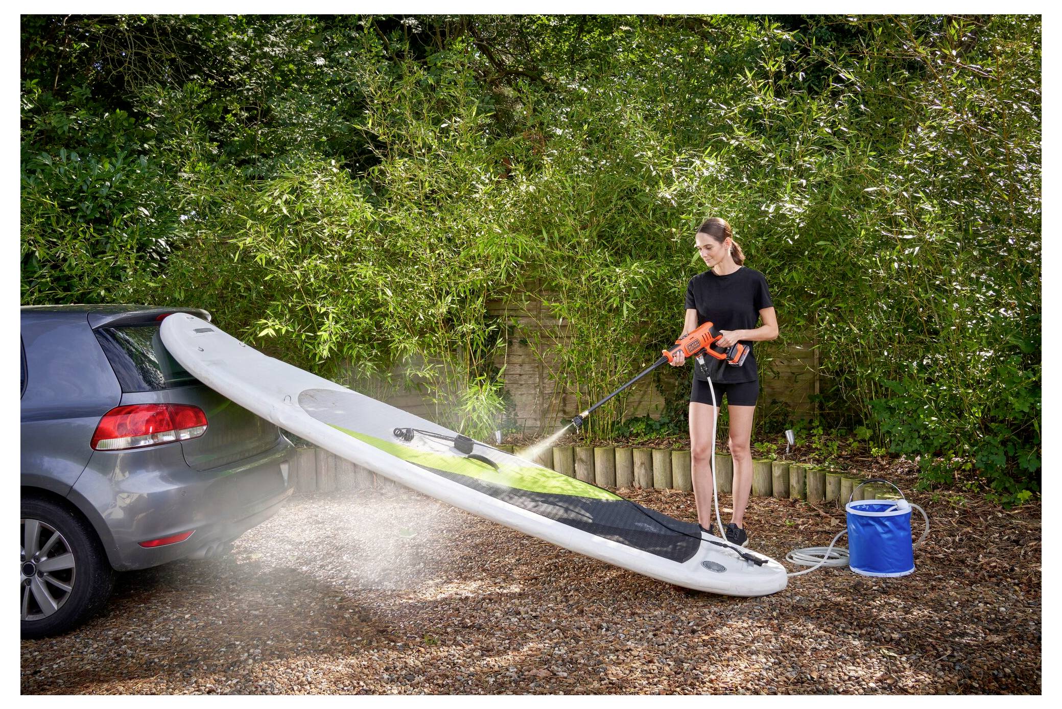 Eine behandschuhte Hand hält den Griff eines orange-schwarzen Elektrowerkzeugs, möglicherweise einer Bohrmaschine, im Freien vor einem unscharfen Rasenhintergrund.