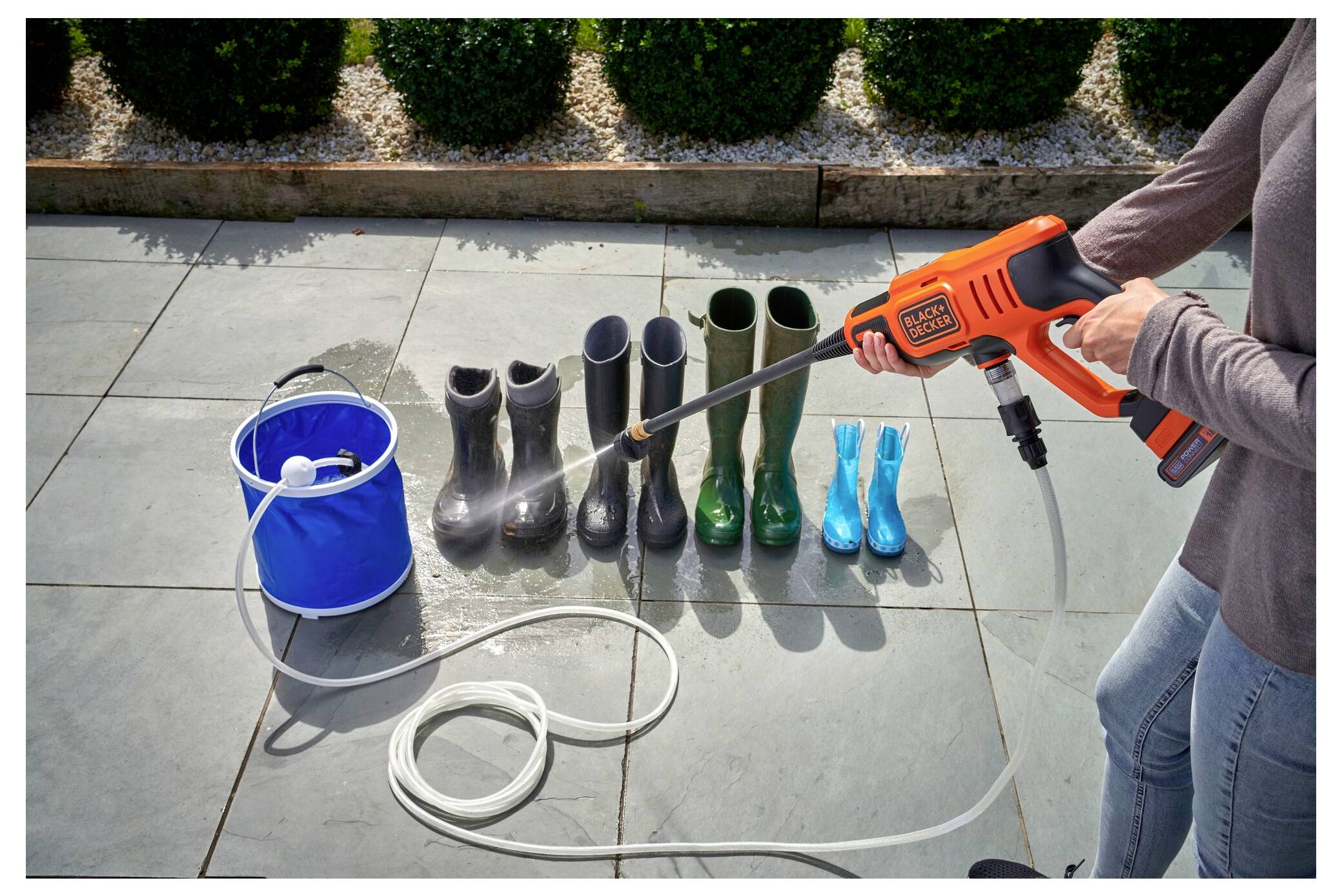 Eine Person nutzt einen orangefarbenen Hochdruckreiniger, um gummierte Stiefel zu reinigen, die auf einer Terrasse aufgereiht sind; ein blauer Eimer voller Wasser steht in der Nähe.