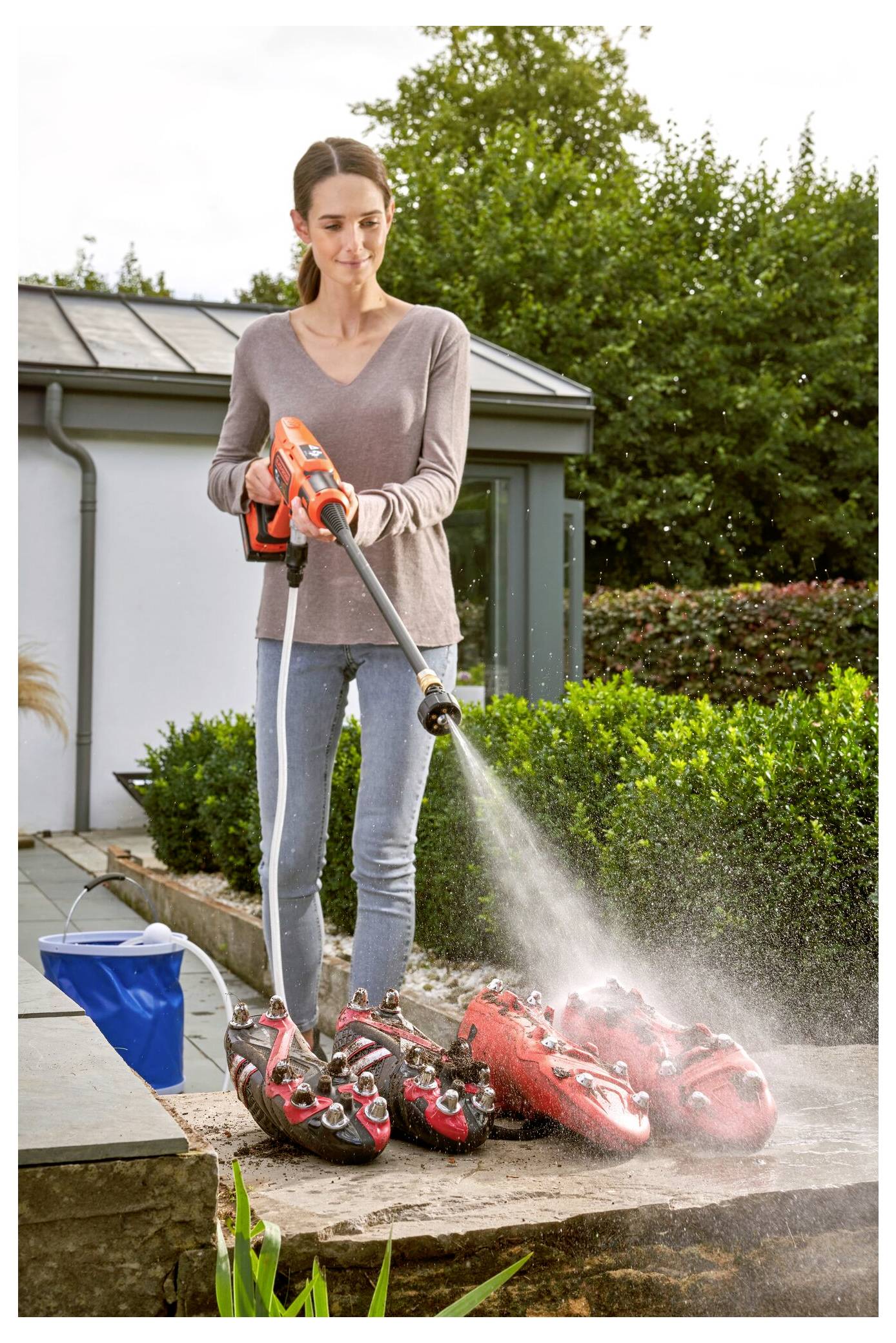 Eine Person benutzt einen Hochdruckreiniger, um rote Fußballschuhe auf einer Steinleiste außerhalb eines Hauses mit Grünflächen im Hintergrund zu reinigen.