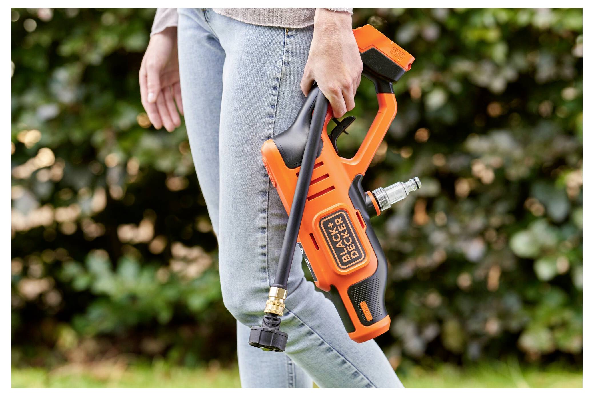 Eine Person hält einen orangefarbenen Black & Decker Hochdruckreiniger und steht im Freien. Das Gerät hat eine lange Düse, die sich für Reinigungsarbeiten eignet.