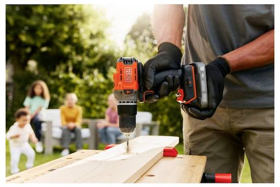 Eine Person mit Handschuhen benutzt einen orangefarbenen kabellosen Bohrer, um ein Loch in ein Holzbrett im Freien zu bohren, während eine Familie im Hintergrund zu sehen ist.