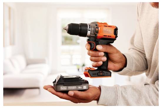 Eine Person hält einen orange-schwarzen kabellosen Bohrer mit einem Ladegerät in einem gut beleuchteten Wohnzimmer.