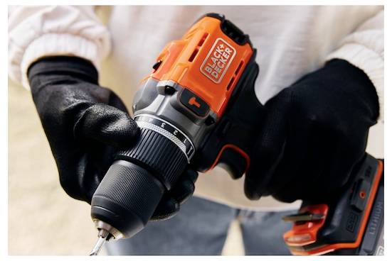 Eine Person, die schwarze Handschuhe trägt, hält einen orange-schwarzen Elektroschrauber der Marke „BLACK+DECKER