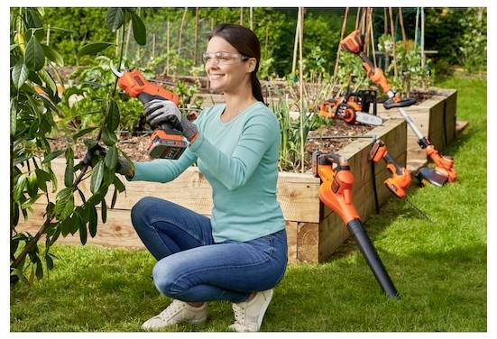 Eine Person, die eine Schutzbrille und Handschuhe trägt, schneidet mit einem kabellosen Heckenschneider eine Pflanze in einem Garten mit erhöhten Holzbeeten.