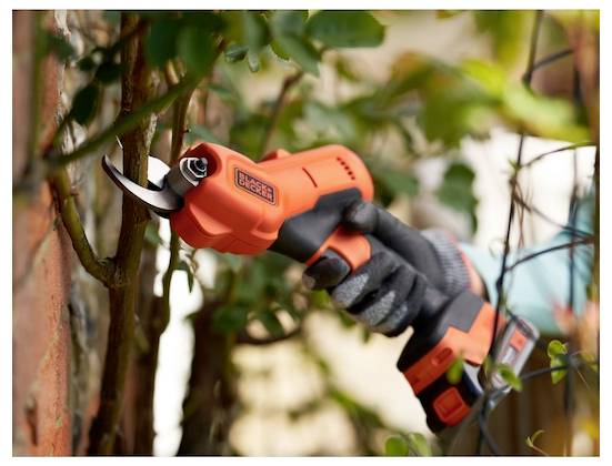 Eine Person nutzt ein elektrisches Schneidewerkzeug, um Äste in einer Gartenumgebung zu schneiden und trägt dabei Schutzhandschuhe.