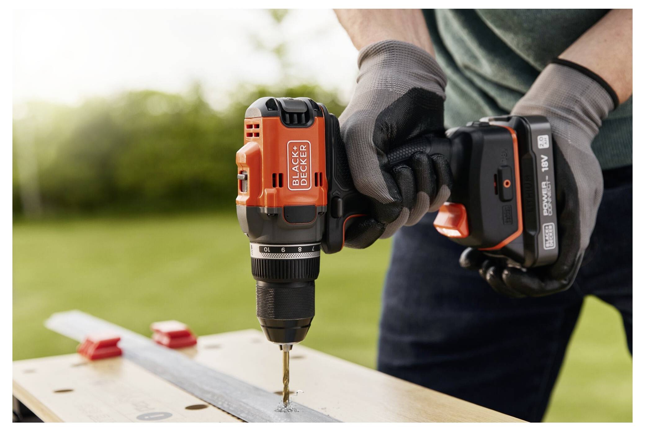 Eine Person mit Handschuhen benutzt einen orange-schwarzen kabellosen Bohrer, um ein Loch in einen Metallstreifen auf einer Werkbank im Freien zu bohren.