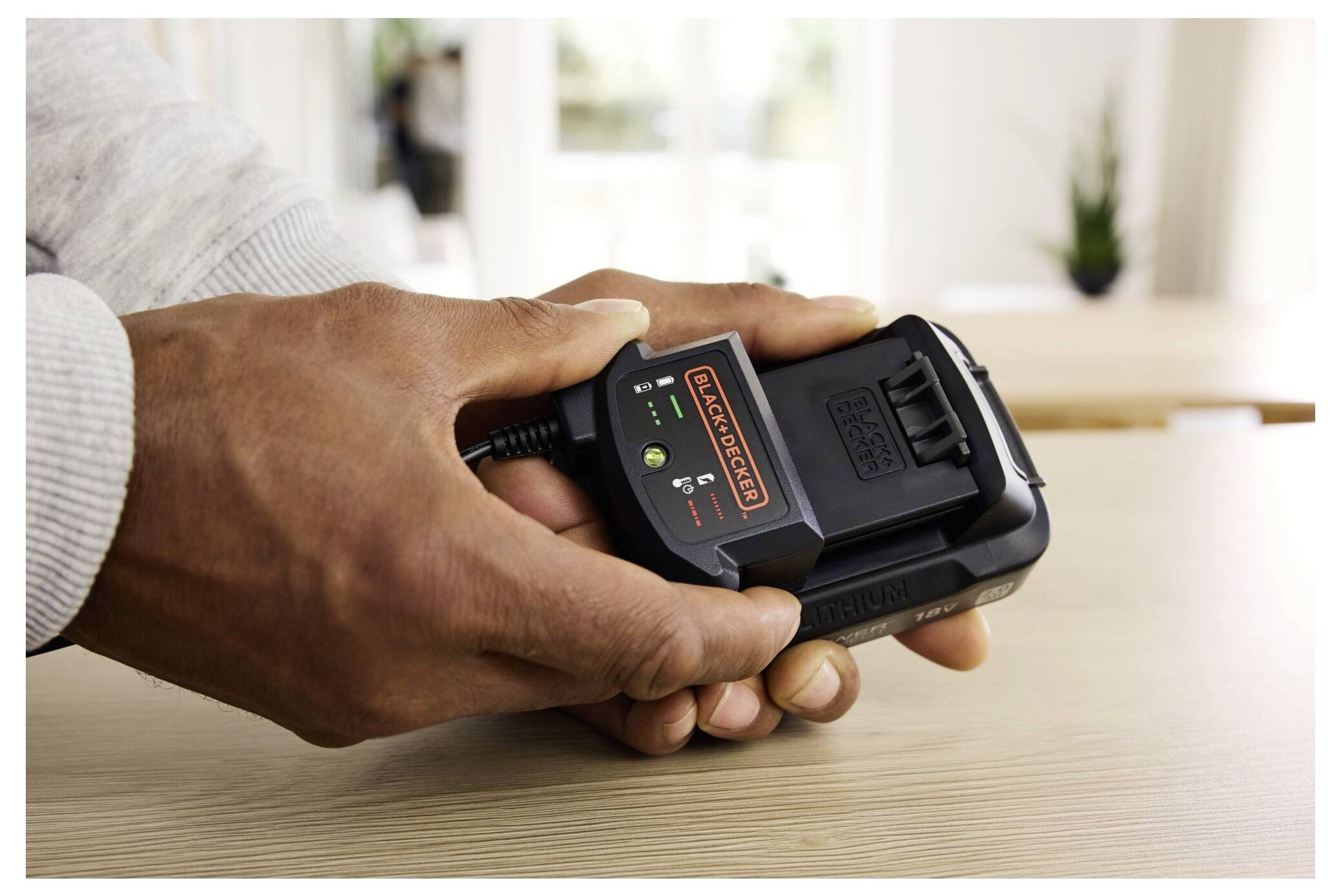 Eine Person hält ein Black+Decker-Batterieladegerät mit Anzeigelichtern, die den Ladestatus anzeigen, auf einem Holztisch.