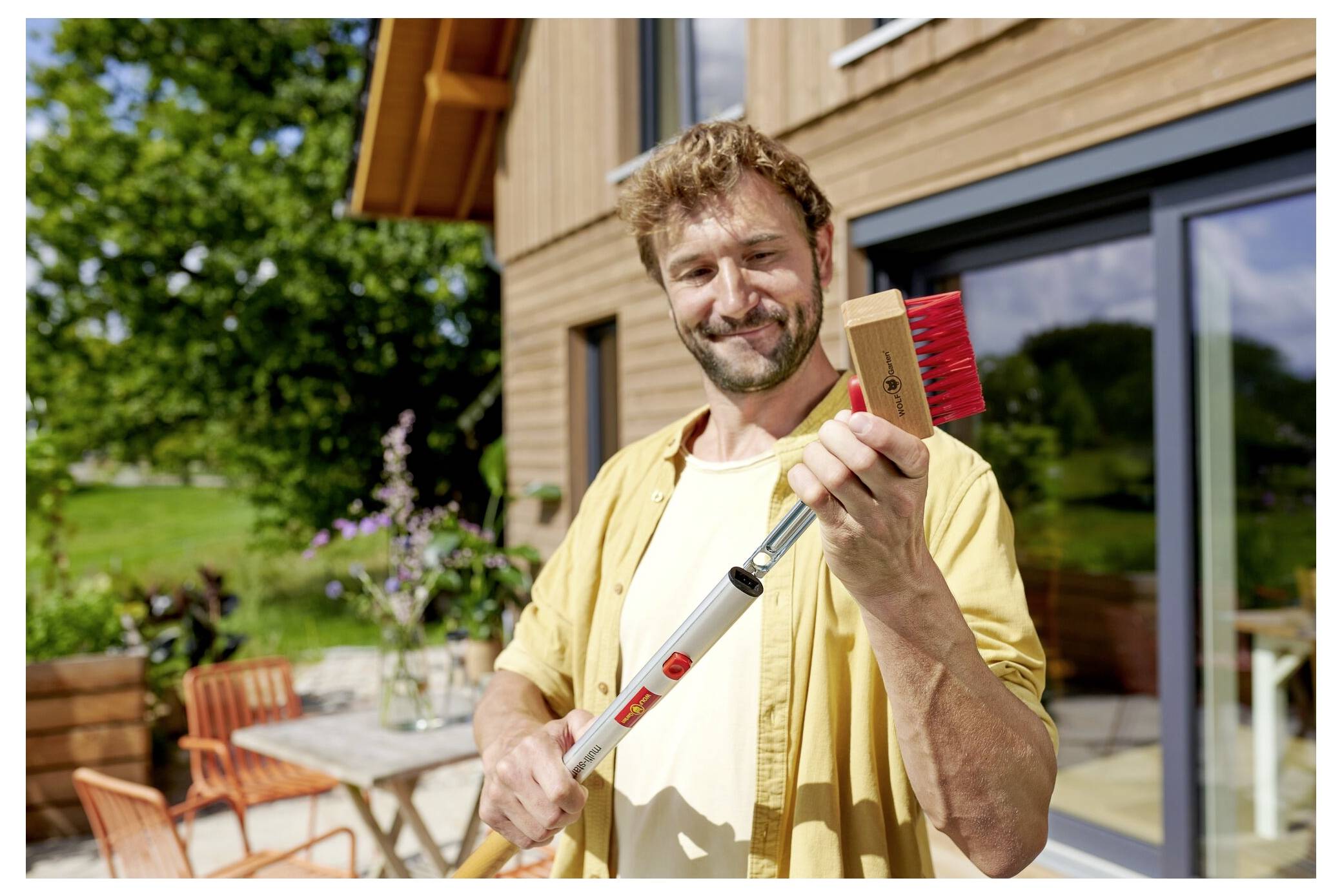 „Eine Person, die lächelt und einen Besen mit roten Borsten hält, steht außerhalb eines Holzhauses mit einem Terrassentisch und Stühlen im Hintergrund.