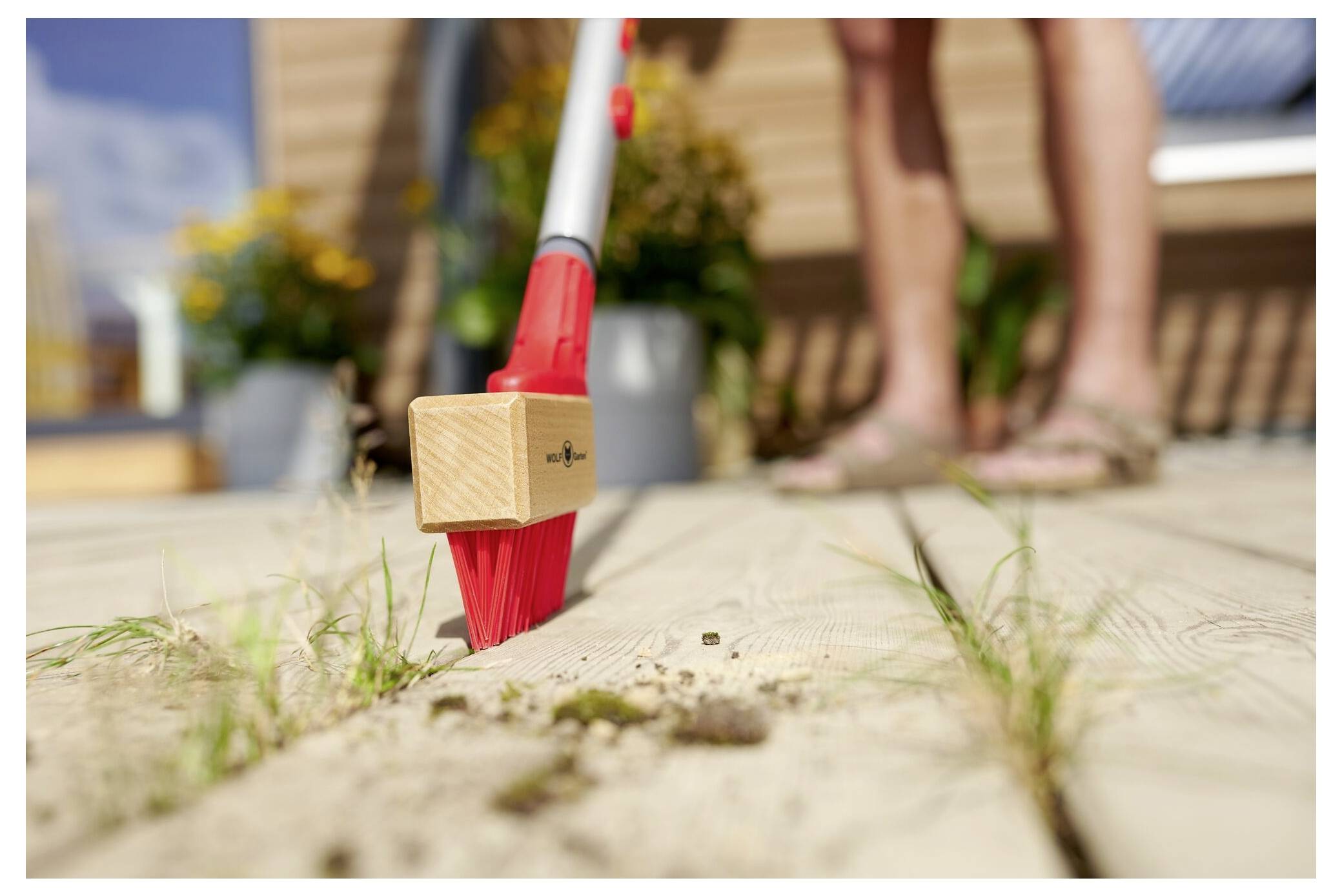 Eine Person benutzt einen roten Besen, um Schmutz und kleine Trümmer von einem Holzdeck zu fegen, mit Pflanzen im Hintergrund, was auf Außenreinigung hindeutet.