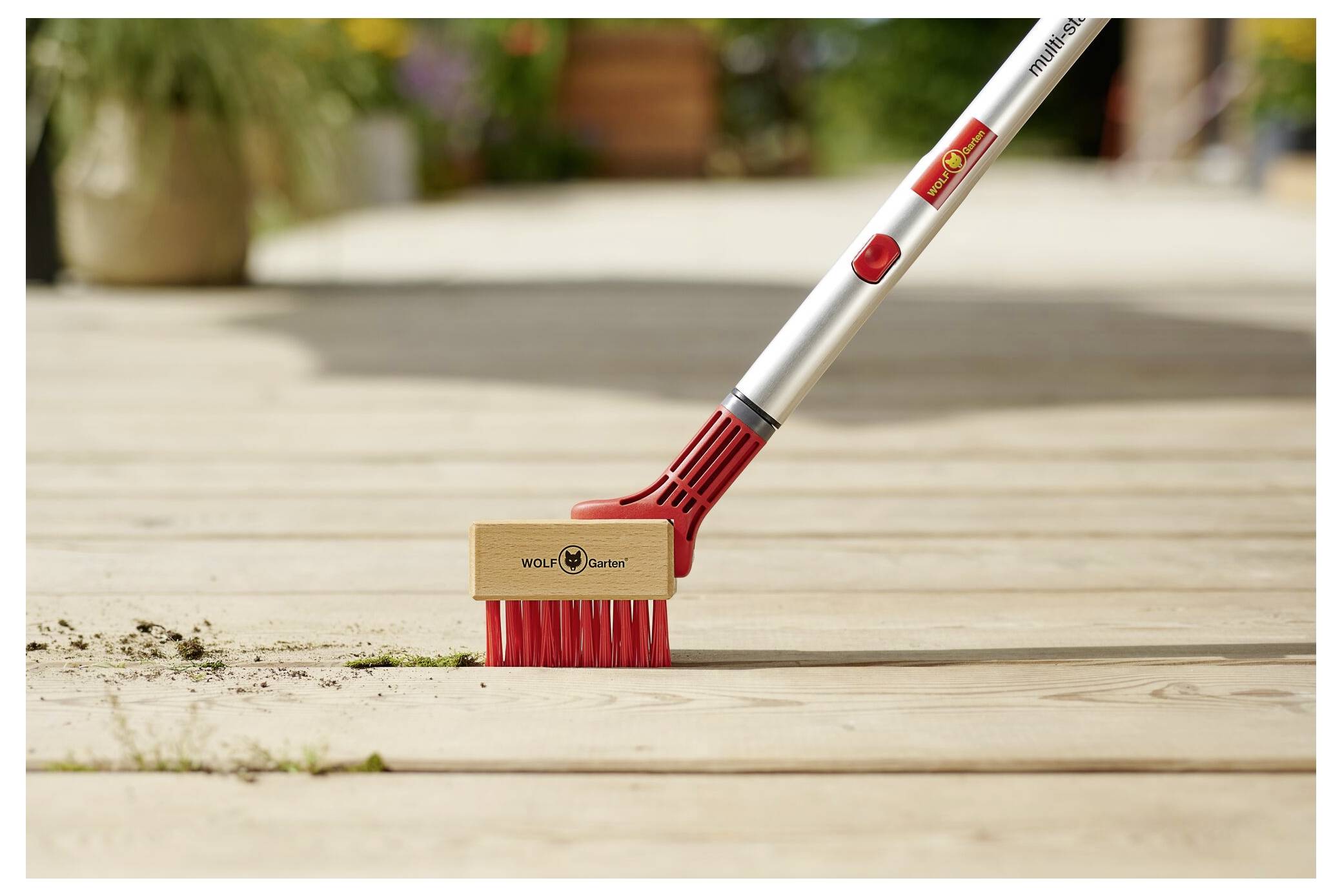 Eine Nahaufnahme einer rot-silbernen Bürste mit Holzkopf, die Schmutz und Unkraut aus den Fugen eines Holzdecks entfernt. Die Bürste hat ein Markenlogo.