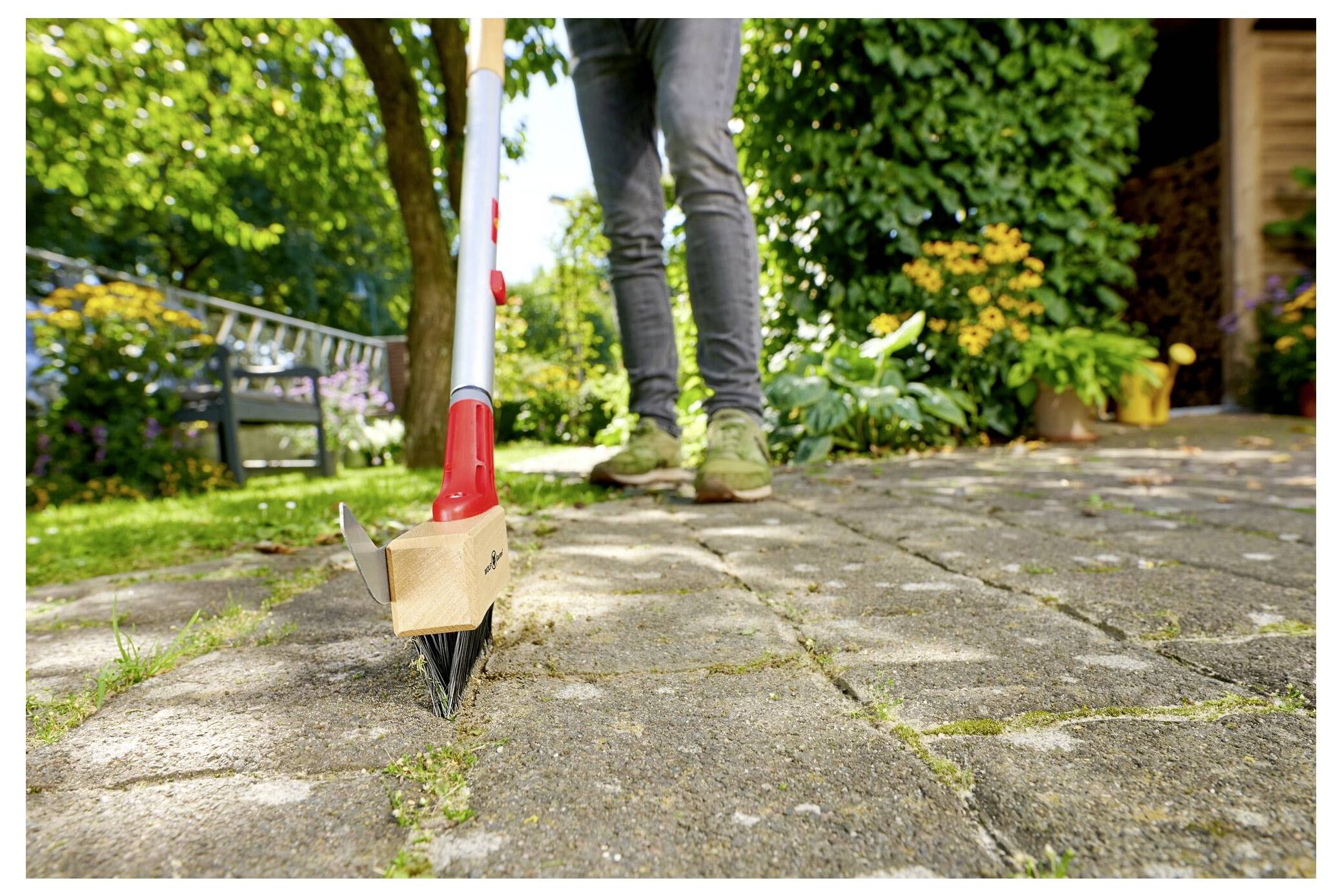 Eine Person, die einen Unkrautvernichter verwendet, um Gras zwischen Terrassensteinen in einem Garten mit Blumen und Grünflächen im Hintergrund zu entfernen.