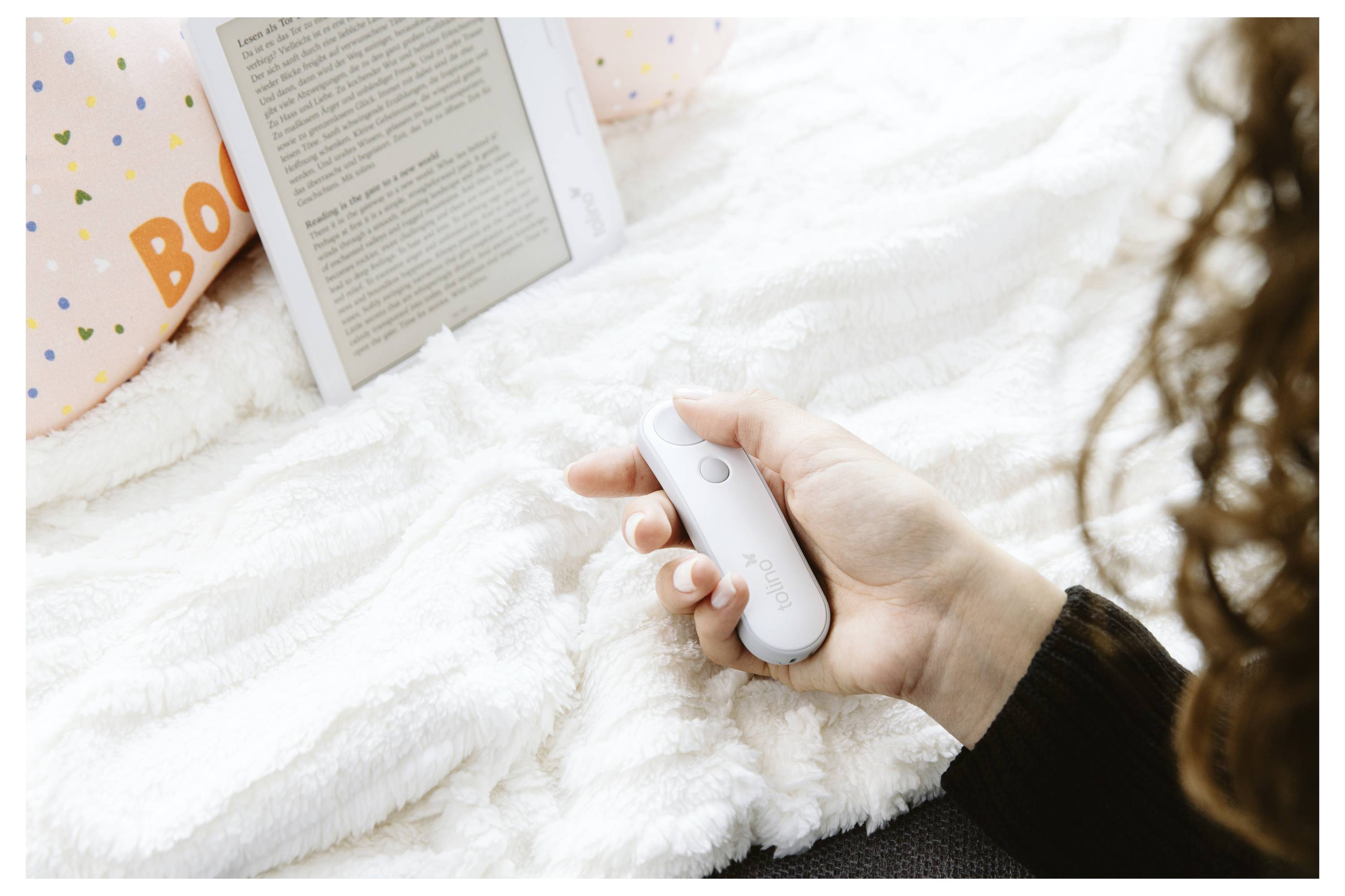 Eine Person hält ein kleines weißes elektronisches Gerät, wahrscheinlich eine Fernbedienung, während ein E-Reader mit angezeigtem Text auf einer weichen, weißen Decke ruht.