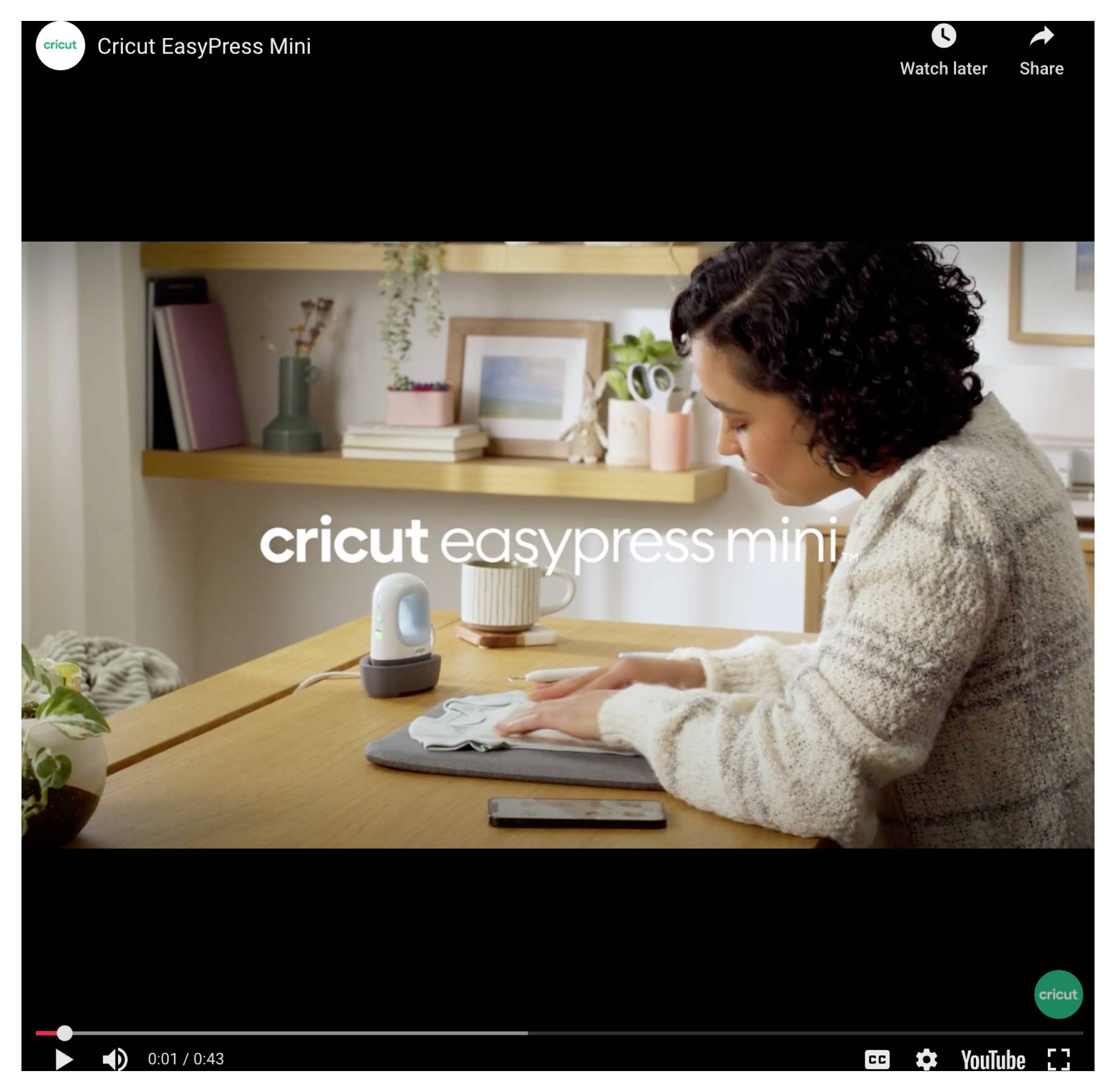 Eine Frau sitzt an einem Tisch und verwendet ein kleines Bügeleisen, um ein Design auf Stoff aufzubringen. Auf dem Bildschirm steht 'Cricut EasyPress Mini'.