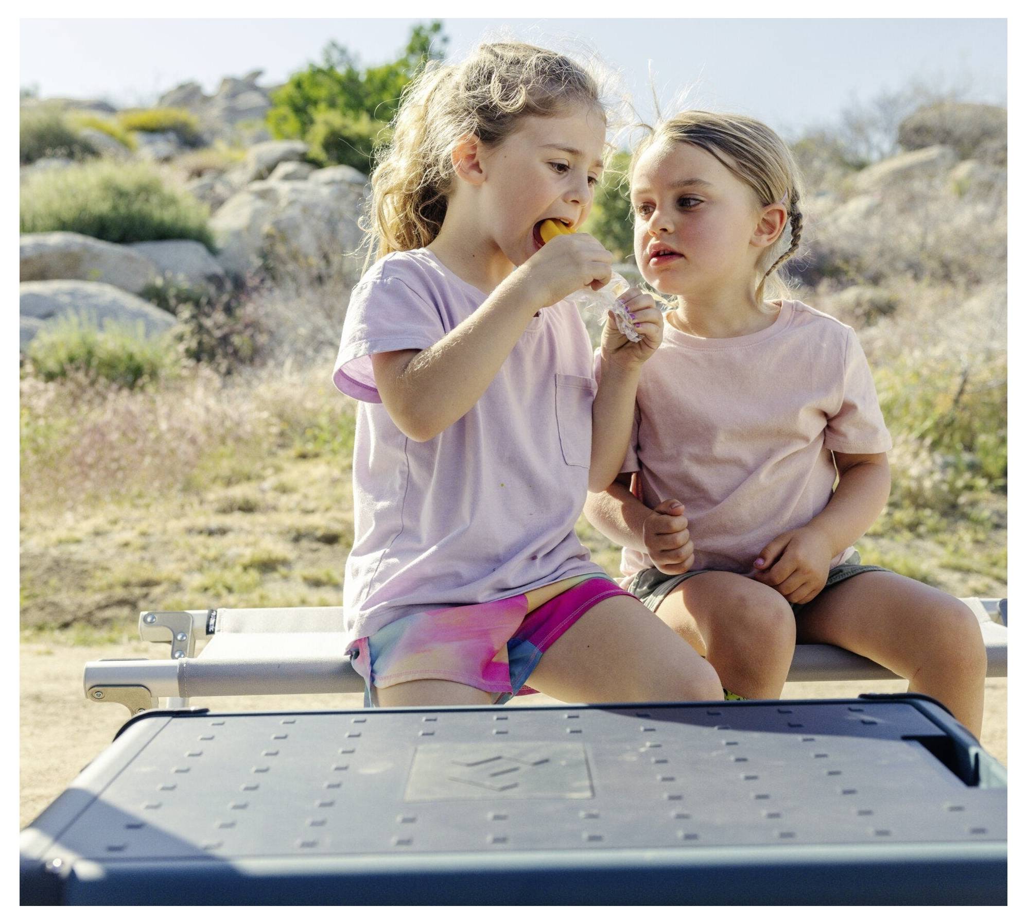 Zwei junge Mädchen sitzen auf einer Bank im Freien und teilen einen Snack in einer sonnigen Felsenlandschaft.