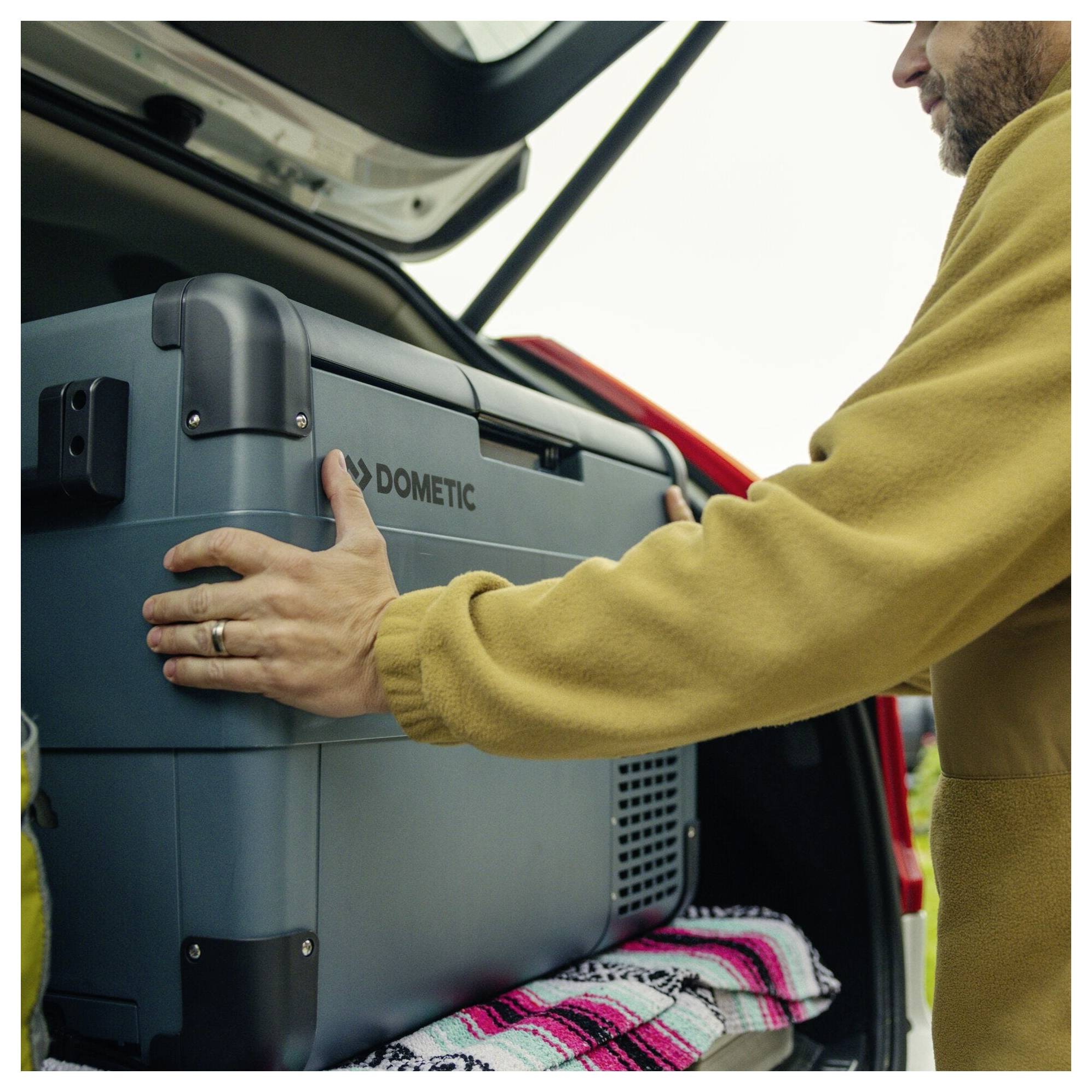 Mann lädt eine Dometic-Kühlbox in den offenen Kofferraum eines Autos, mit einer Decke darunter.