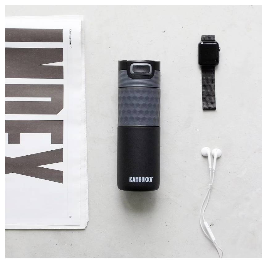 Ein schwarze Kambukka-Flasche, eine zusammengerollte Zeitung, eine Apple Watch und weiße Kopfhörer auf einer weißen Oberfläche angeordnet.