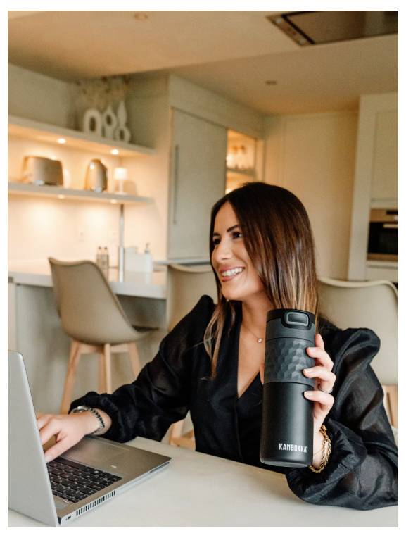 Eine Frau in einer Küche hält eine Thermoskanne und lächelt auf einen Laptop-Bildschirm. Apps und Dekoration sind auf einem Regal im Hintergrund sichtbar.