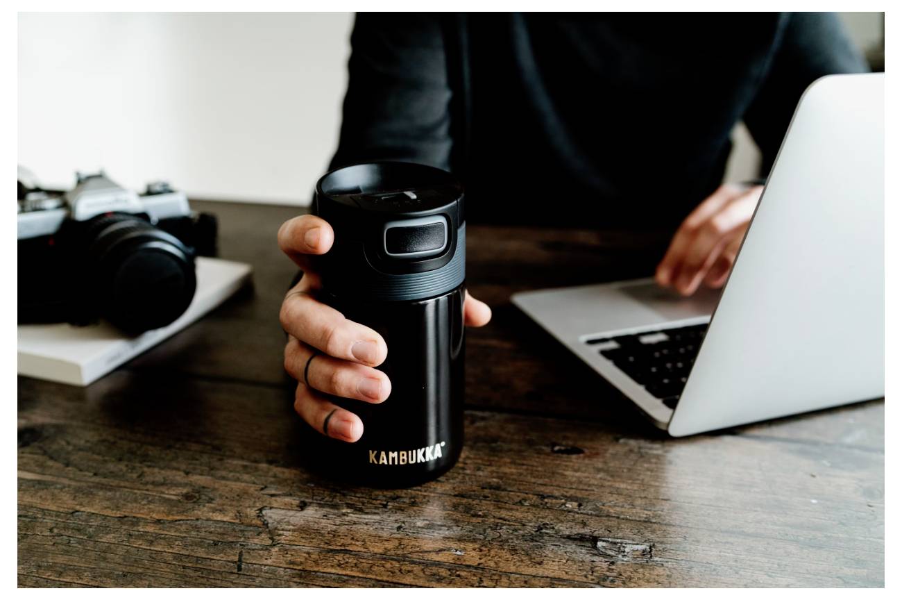 Eine Person sitzt an einem Holztisch und hält einen schwarzen Reisebecher mit der Aufschrift „Kambukka