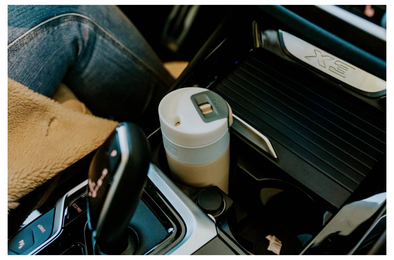 Eine Reisekaffeetasse in der Getränkehalterung eines Autos neben dem Schalthebel, mit einer Person in Jeans und beigem Stoff, die daneben sitzt.