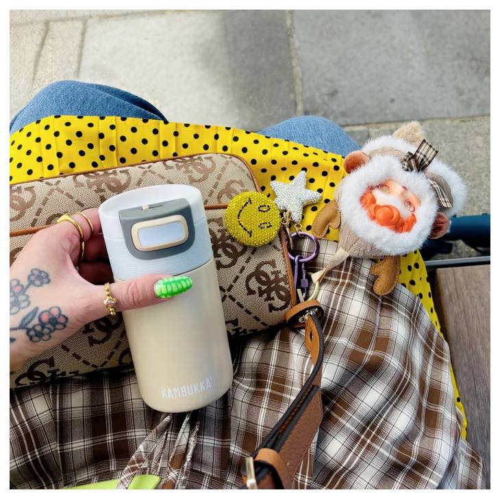 Eine Person mit einer tätowierten Hand hält eine beige Kambukka-Flasche und eine gemusterte Tasche, die mit verschiedenen Schlüsselanhängern verziert ist, einschließlich eines Stoff-Pilzes.