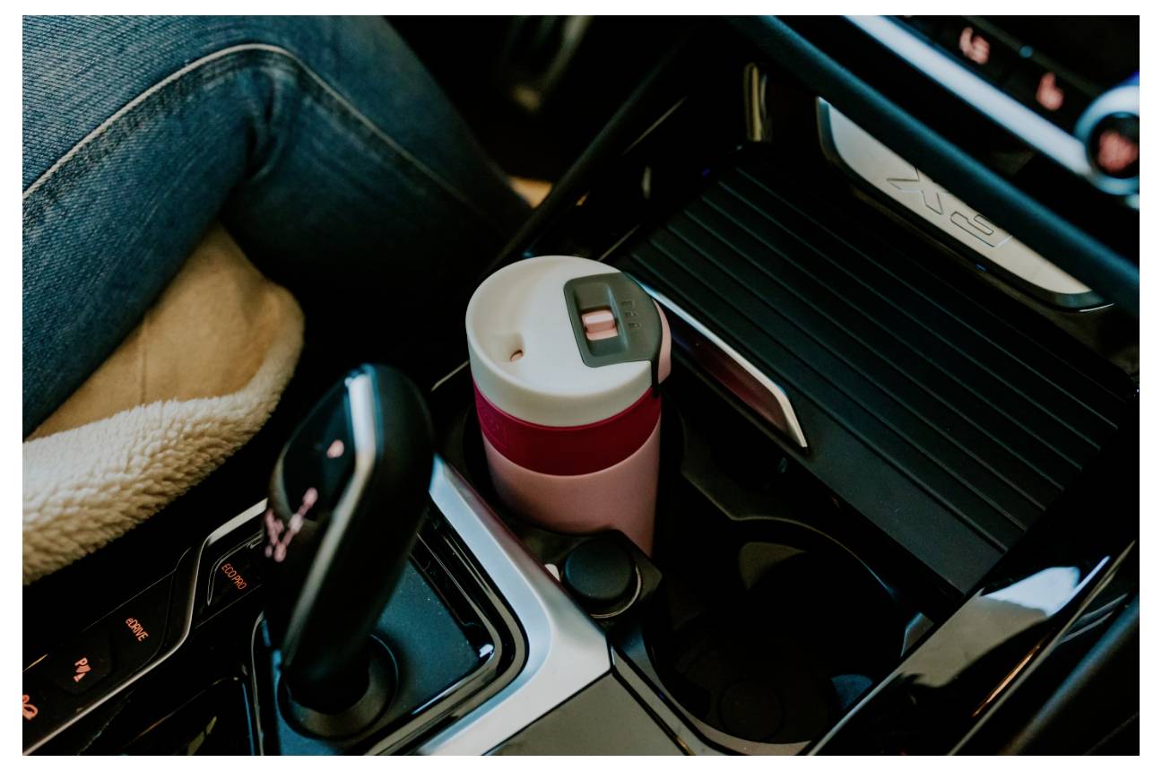 Eine rosa Reisebecher in der Mittelkonsole eines Autos neben einem Schaltknüppel, mit einer Person, die auf der linken Seite sitzt und Jeans trägt, und einem beigen Kissen, das sichtbar ist.