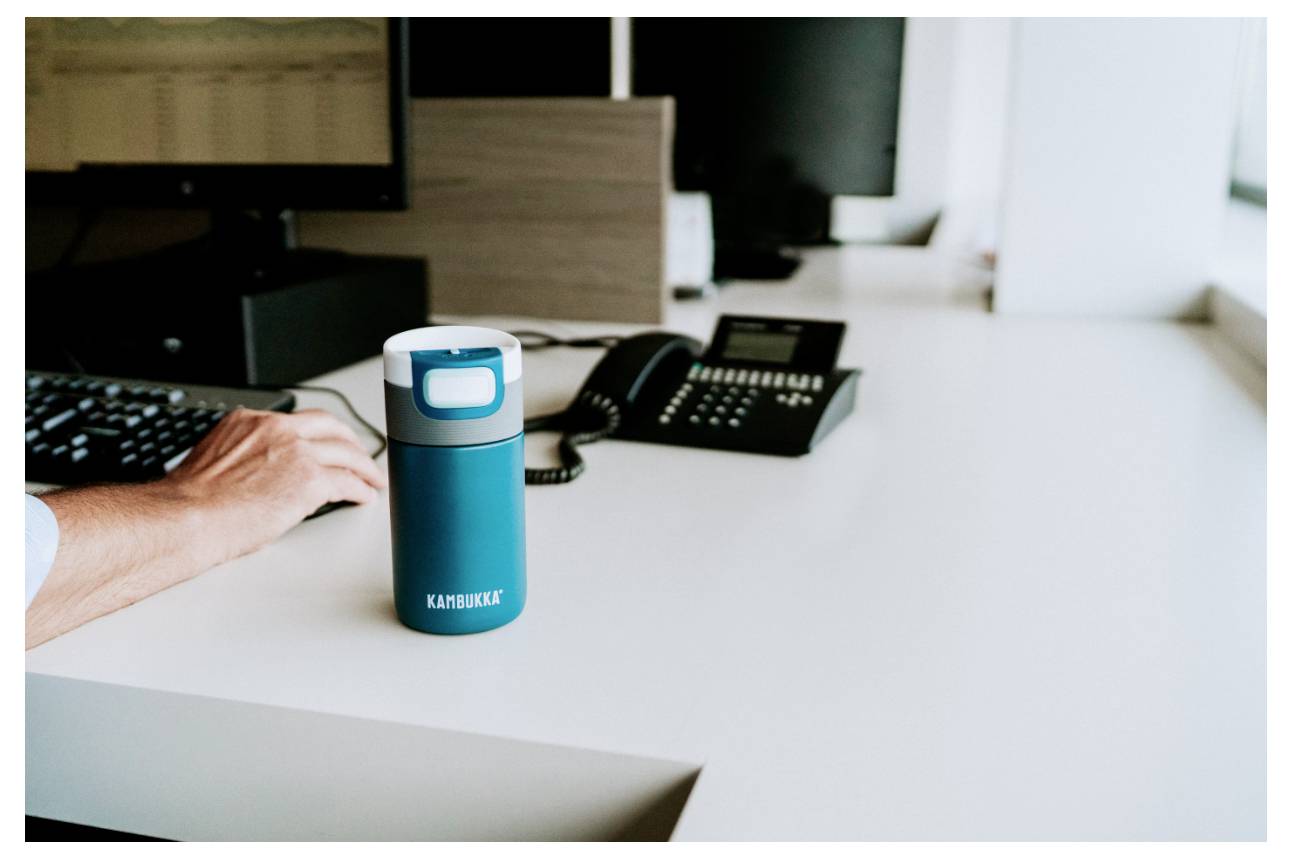 Eine Person, die neben einer blauen Reisekaffeetasse an einem weißen Schreibtisch an einer Tastatur arbeitet, mit einem Telefon und Monitoren im Hintergrund.