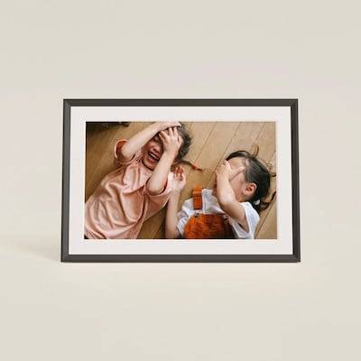 Ein gerahmtes Foto von zwei Kindern, die auf einem Holzboden liegen, lächeln und Verstecken spielen, wobei ihre Hände ihre Gesichter bedecken.
