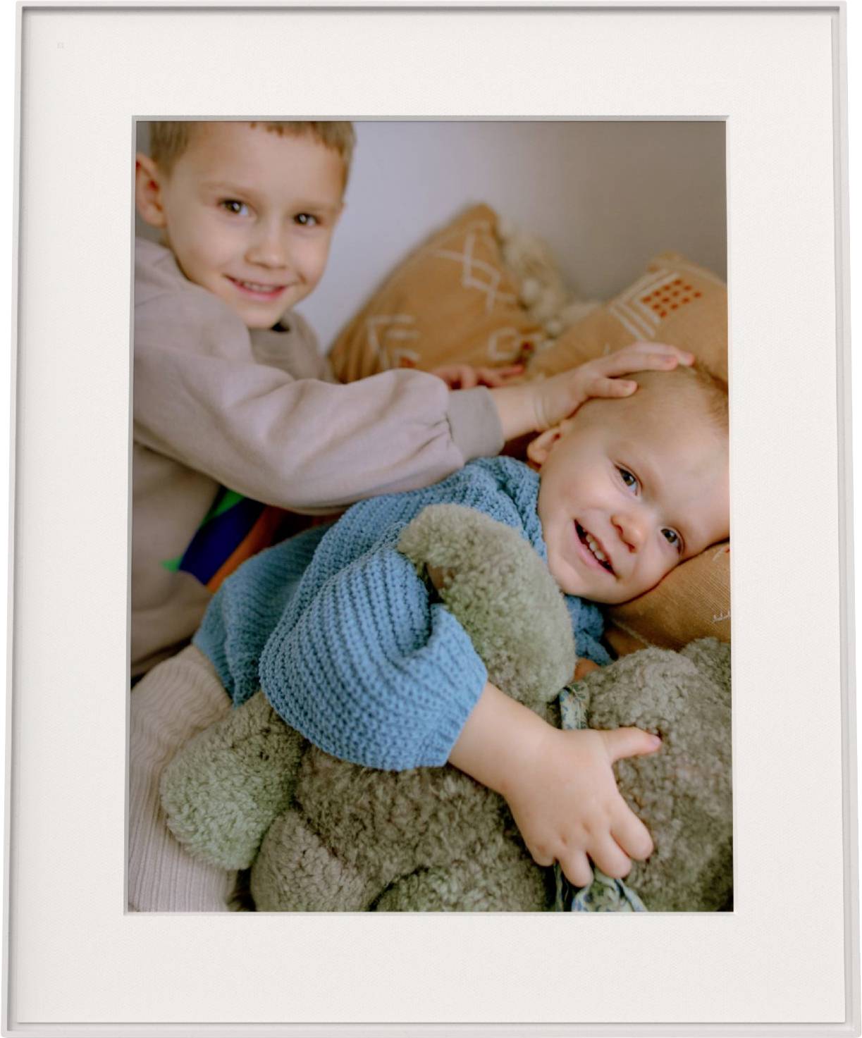 Zwei kleine Kinder, eines lächelnd und eine Kuscheltier umarmend, das andere spielerisch den Kopf des ersten Kindes tätschelnd, während beide auf einem Bett sitzen.