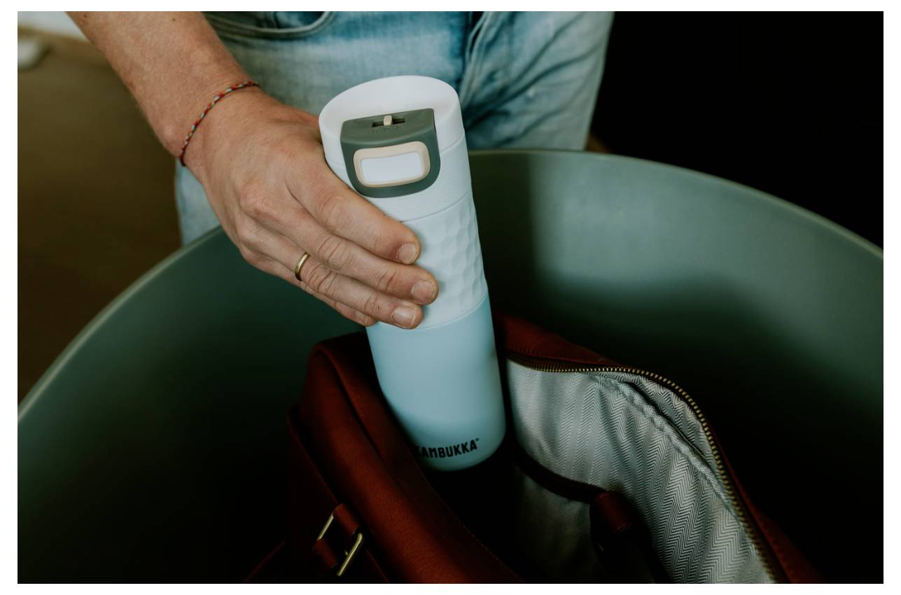 Eine Person legt eine blaugrüne Wasserflasche in eine braune Tasche, die ein Logo einer Hydratationsmarke zur Schau stellt, in einer Nahaufnahme.